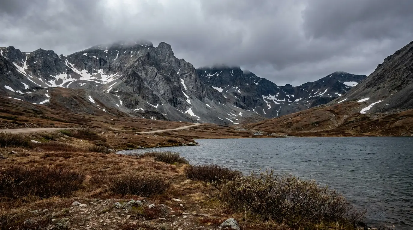 A treeless alpine lake at a windswept mountain pass ringed by bare rocky peaks with lingering snow patches, tundra and low shrubs in the foreground, dramatic cold cloudy light, Summit Lake Stone Mountain Provincial Park British Columbia, ultrarealistic photograph representative of the region