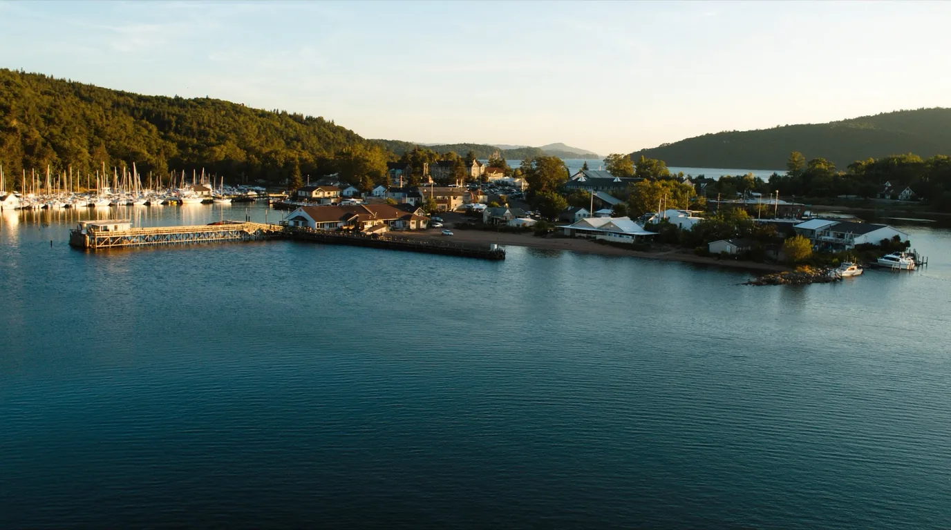 A small Great Lakes harbour town waterfront with moored sailboats and fishing boats, clapboard buildings on a hillside above the docks, cold blue Lake Superior beyond, Bayfield Wisconsin, ultrarealistic photograph representative of the region