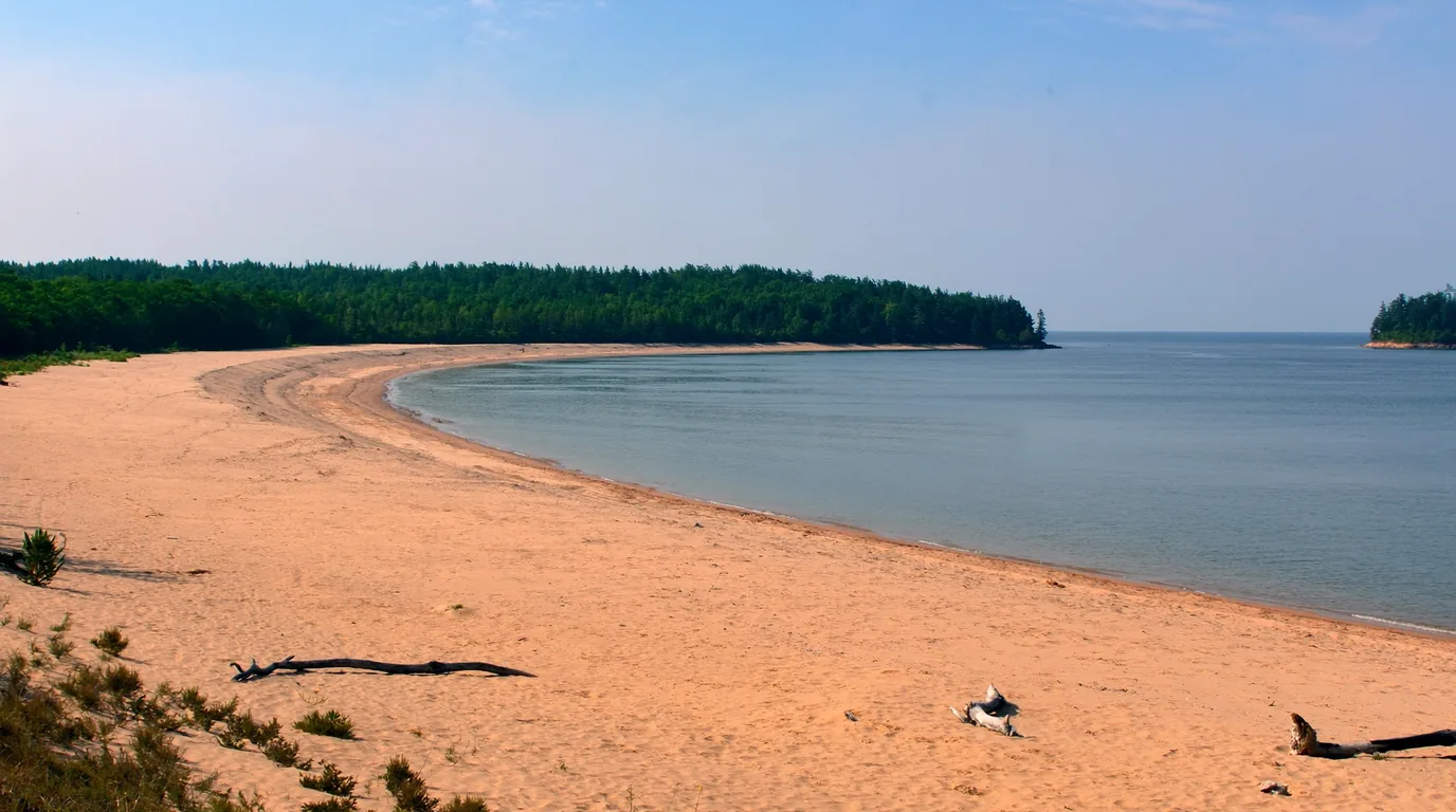 A long empty pale sand beach on a Lake Superior island with clear shallow turquoise water, driftwood at the treeline and dark spruce forest behind, nobody on the beach, Apostle Islands Wisconsin, ultrarealistic photograph representative of the region