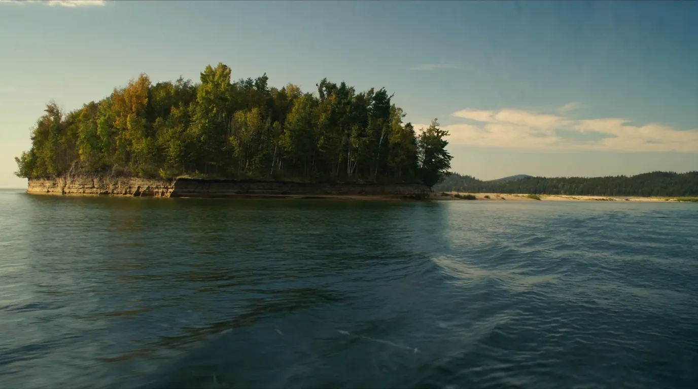 A low wooded island seen across open cold blue-grey Lake Superior water under a wide pale sky, dark spruce and birch on its shore, no buildings anywhere, Apostle Islands Wisconsin, ultrarealistic photograph representative of the region