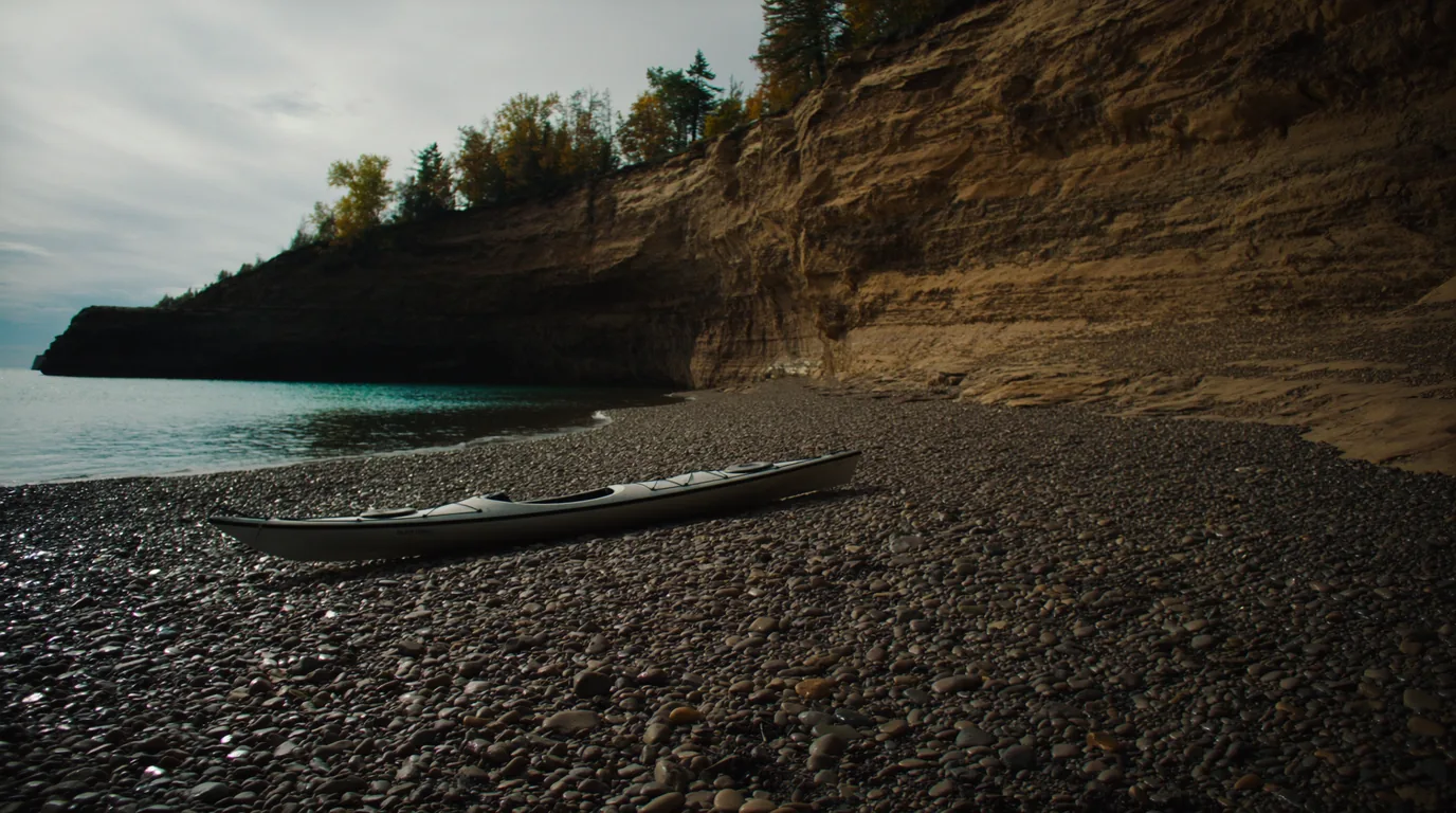A sea kayak drawn up on a cobble shore beneath layered red sandstone cliffs on Lake Superior, paddle across the deck, calm clear cold water, Apostle Islands Wisconsin, ultrarealistic photograph representative of the region
