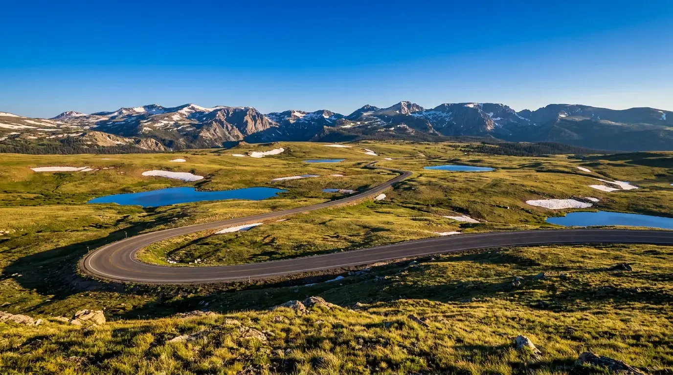 A high alpine highway switchbacking across a treeless plateau of tundra, lingering snowfields and small alpine lakes above 10,000 feet with jagged granite peaks on the horizon under warm morning light, Beartooth Plateau Montana Wyoming, ultrarealistic photograph representative of the region