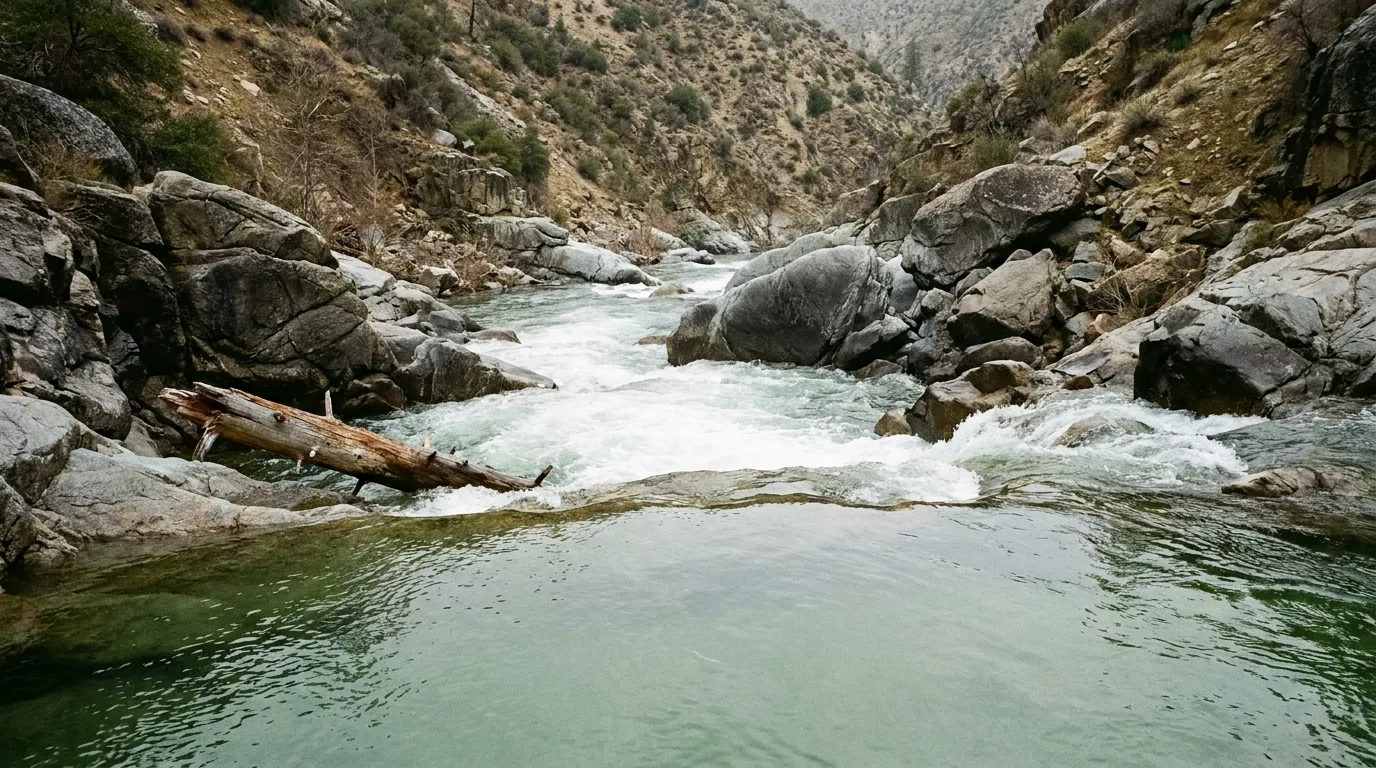 A deceptively calm glassy green pool on a mountain river in the foreground sliding over a lip into violent whitewater and undercut granite boulders beyond, a fallen pine trunk half-submerged at the edge, steep dry canyon walls, flat overcast light, Kern River southern Sierra Nevada California, ultrarealistic photograph representative of the region