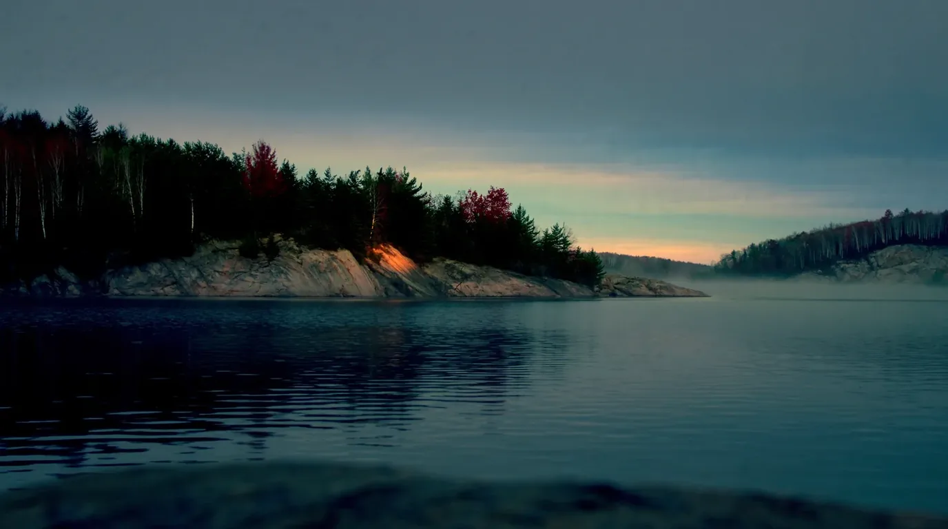A still Boundary Waters lake at dawn with mist on the water, low granite outcrops and dark spruce and pine along the shoreline, a canoe drawn up on the rock, northern Minnesota, ultrarealistic photograph representative of the region