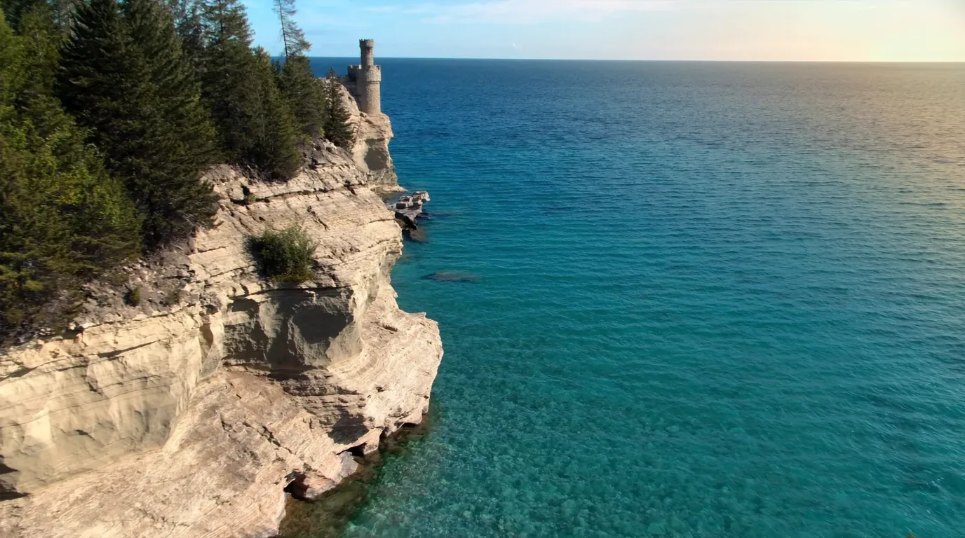 A detached sandstone pillar standing separate from the shoreline cliff with a single white pine growing on its flat top and roots running horizontally through the air back to the mainland, clear Lake Superior water below, Chapel Rock Michigan, ultrarealistic photograph representative of the region