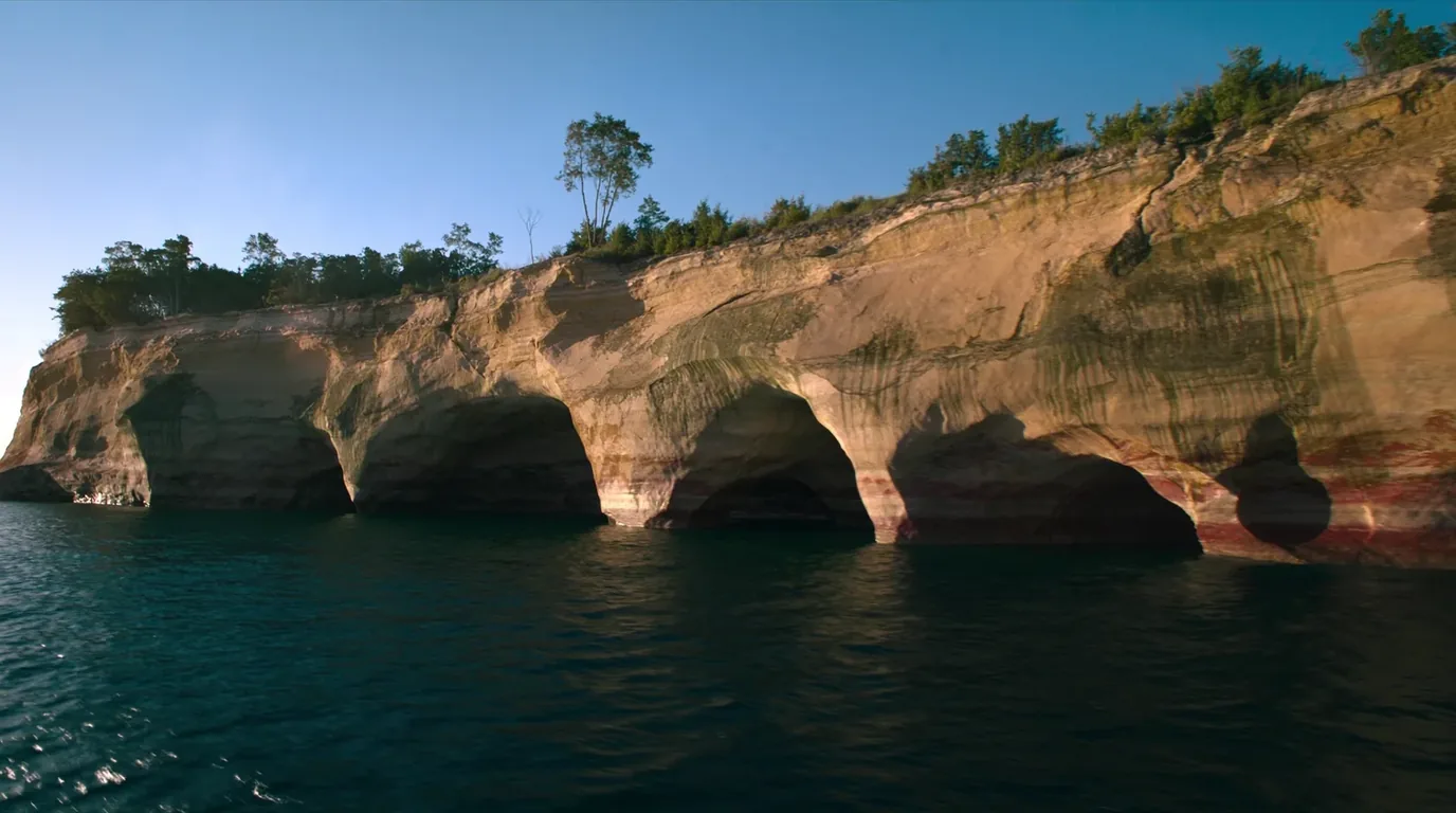 Layered sandstone cliffs streaked with mineral stains of orange, green and black rising straight out of the clear cold turquoise water of Lake Superior, dark conifer forest along the clifftop, Pictured Rocks Michigan, ultrarealistic photograph representative of the region