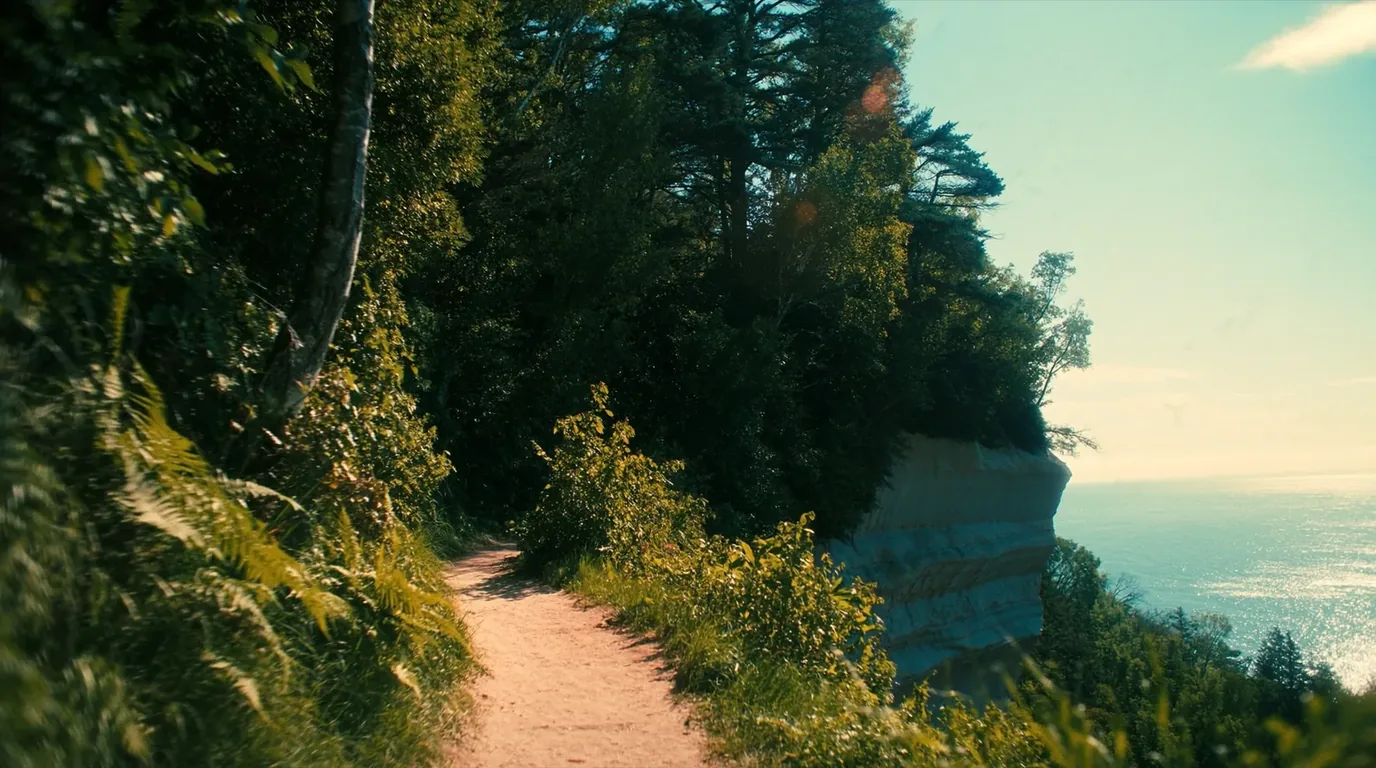 A narrow footpath running along the top of high layered sandstone cliffs above the clear cold water of Lake Superior with birch and conifer forest inland and an open horizon, North Country Trail Michigan, ultrarealistic photograph representative of the region