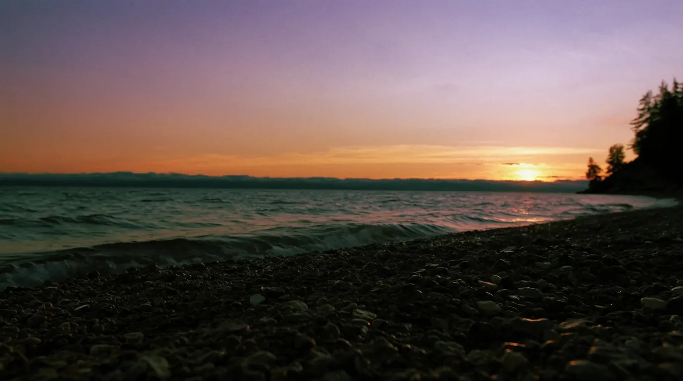 Sunset over Lake Superior seen from a beach of smooth rounded cobbles with warm low light on cold grey-blue water and dark conifers along the shore, Michigan Upper Peninsula, ultrarealistic photograph representative of the region