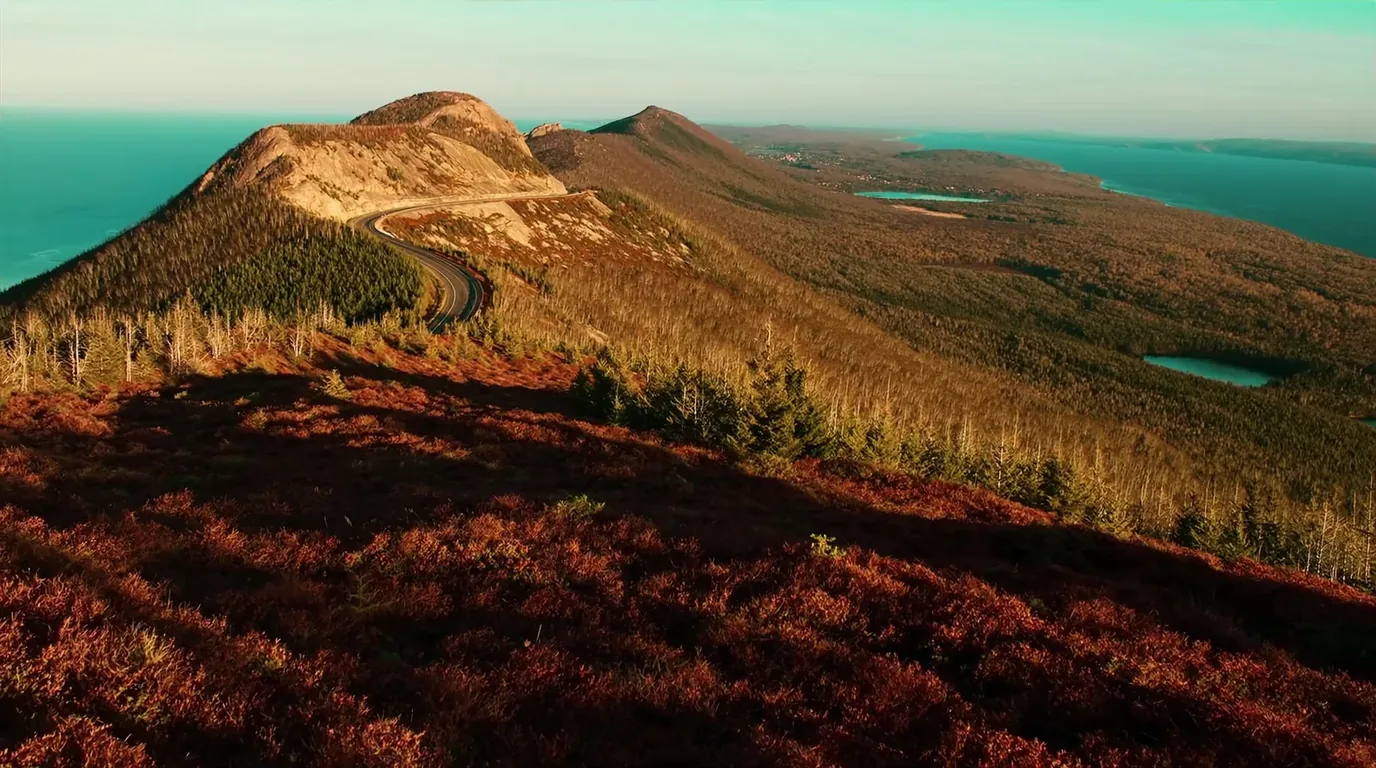 The view from a high ridge road above Copper Harbor with Lake Superior visible on both sides, forested hills falling away and a wide horizon of blue water under evening light, Keweenaw Michigan, ultrarealistic photograph representative of the region