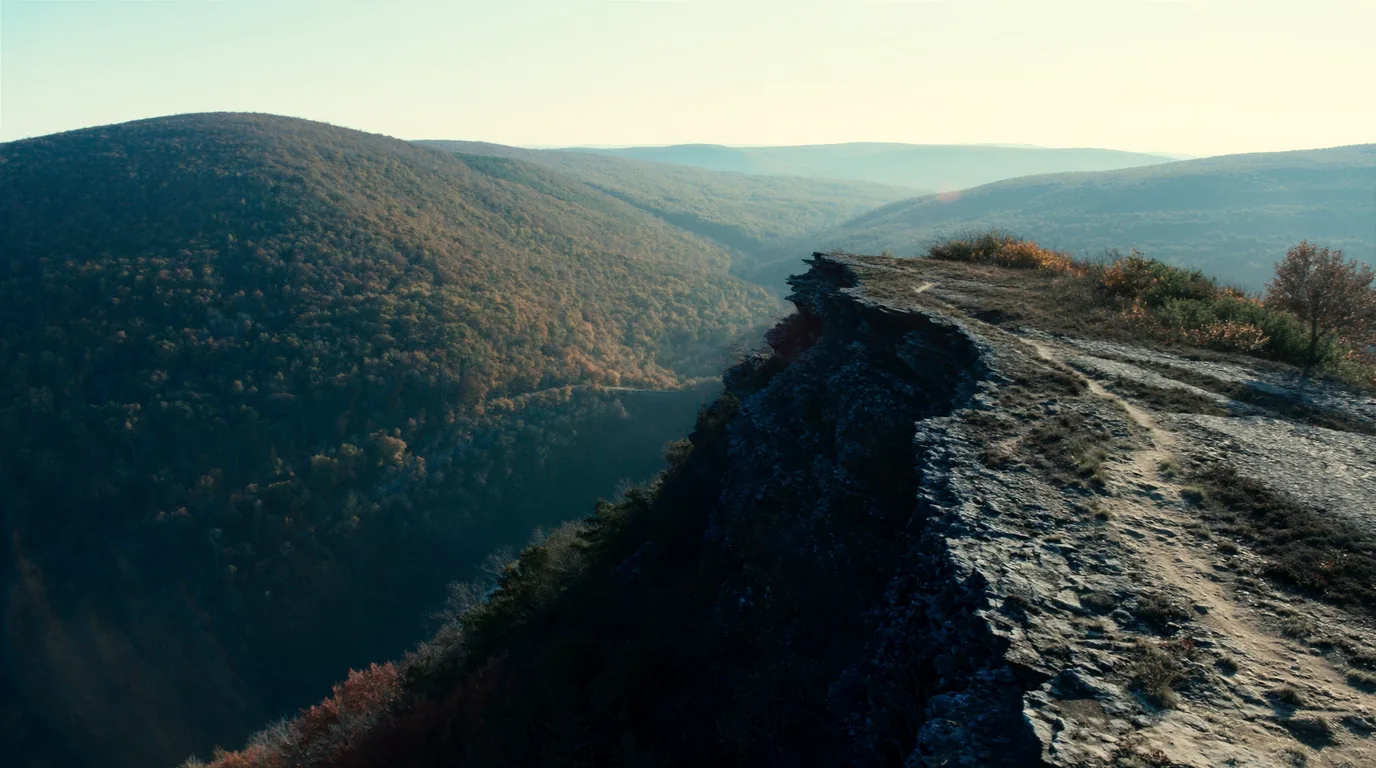 A narrow trail running along an open rocky ridge crest high above a forested valley with a long dark lake below, layered ridges to the horizon, autumn colour in the hardwoods, Porcupine Mountains Michigan, ultrarealistic photograph representative of the region