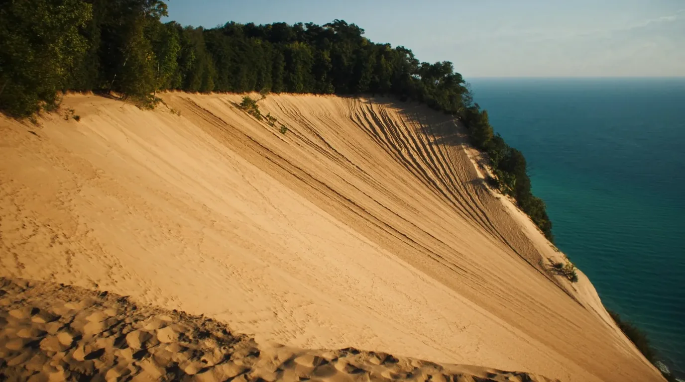 A vast perched sand dune standing four hundred feet above the deep blue water of Lake Michigan with pale sand falling steeply to the shoreline and beech-maple forest inland, Sleeping Bear Dunes Michigan, ultrarealistic photograph representative of the region