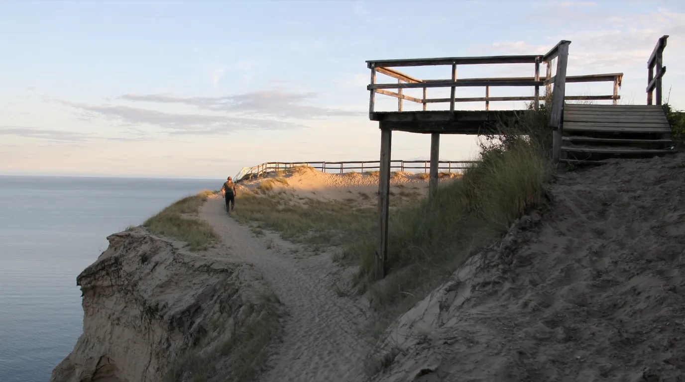 A high dune overlook platform above Lake Michigan at evening with pale sand falling steeply away four hundred and fifty feet to deep blue water, layered lake shelves visible and an open horizon, Sleeping Bear Dunes Michigan, ultrarealistic photograph representative of the region