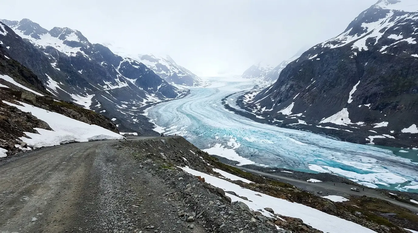 An immense river of blue-white glacial ice curving between dark mountain ridges seen from a high gravel road viewpoint looking down its full length, patches of summer snow, no guardrail, Salmon Glacier above Hyder Alaska, ultrarealistic photograph representative of the region
