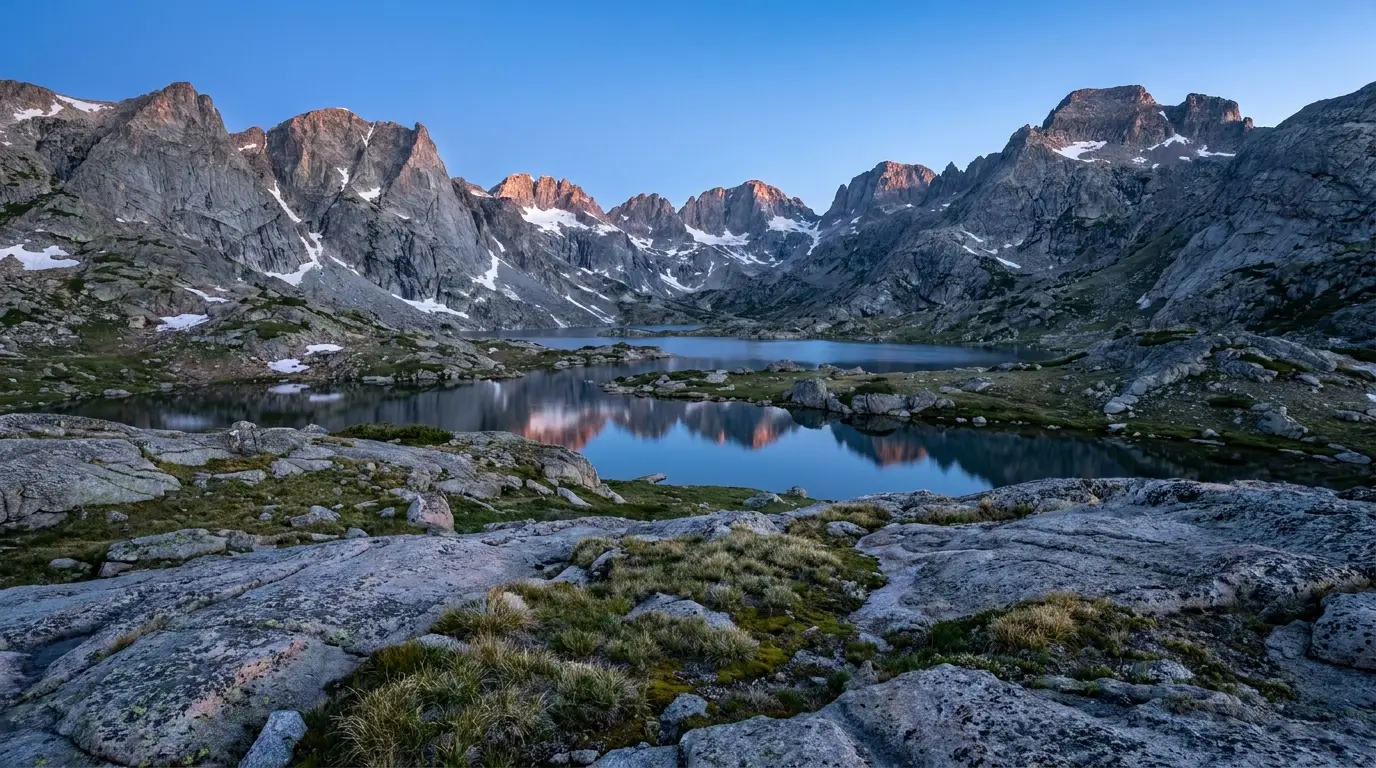 A vast amphitheatre of bare grey granite peaks rising above a chain of still alpine lakes at first light, snow lingering in the high couloirs, granite slabs in the foreground, cold clean high-altitude morning light, Titcomb Basin in the Wind River Range Wyoming, ultrarealistic photograph representative of the region